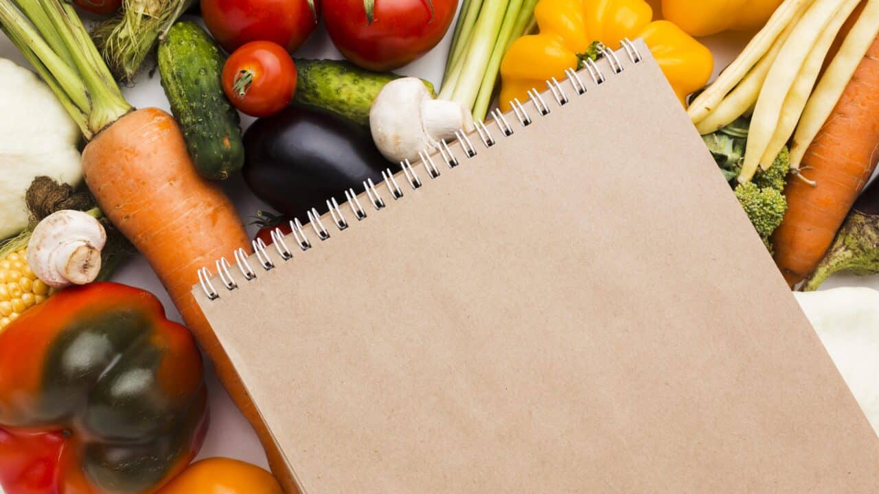Flat lay of fresh vegetables next to a blank notepad, representing a healthy grocery list for meal prep