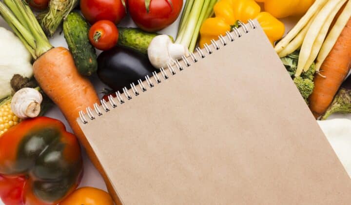 Flat lay of fresh vegetables next to a blank notepad, representing a healthy grocery list for meal prep