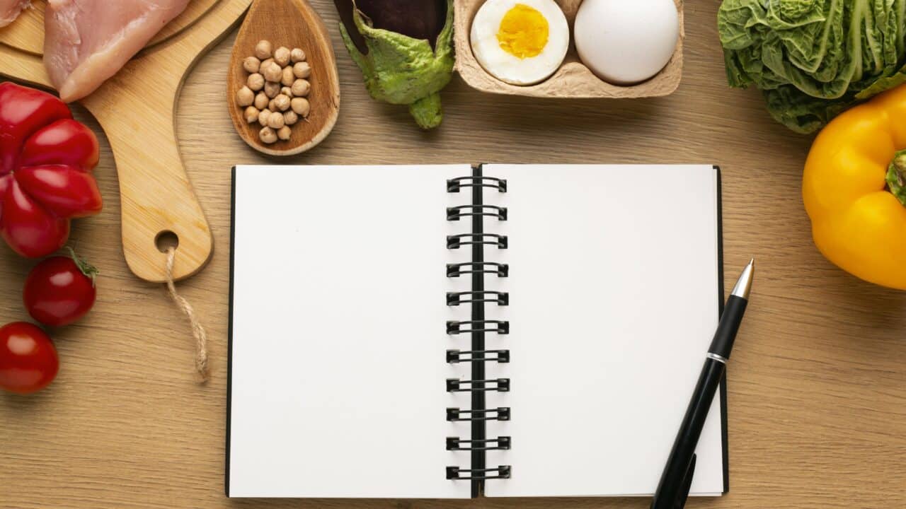 Flat-lay of a meal planning notepad with fresh ingredients arranged around it, overhead view on a neutral background