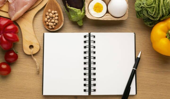 Flat-lay of a meal planning notepad with fresh ingredients arranged around it, overhead view on a neutral background