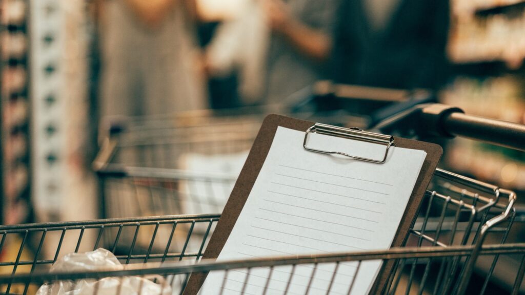 A grocery list on a clipboard resting in a shopping cart in a supermarket aisle