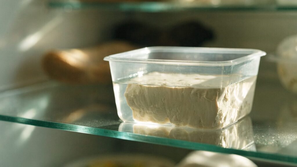 A block of tofu submerged in water inside a clear container in the refrigerator