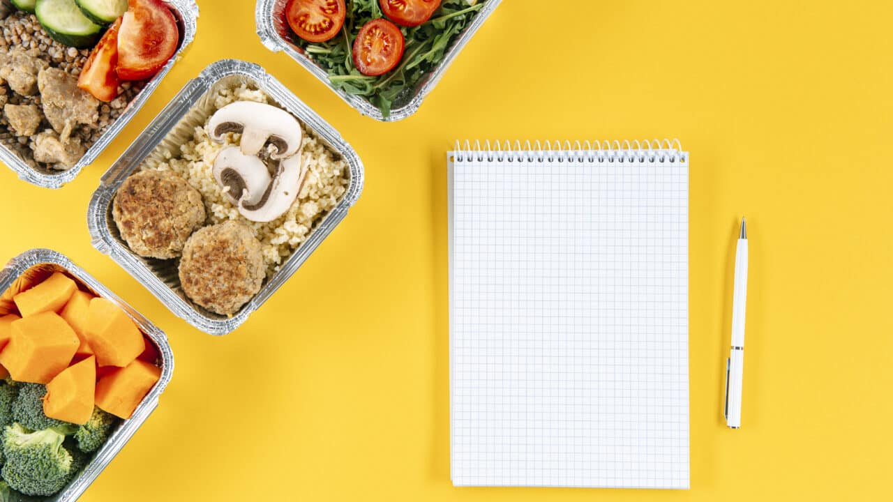 A blank spiral notebook and pen next to foil containers of cooked meal prep food on a yellow background