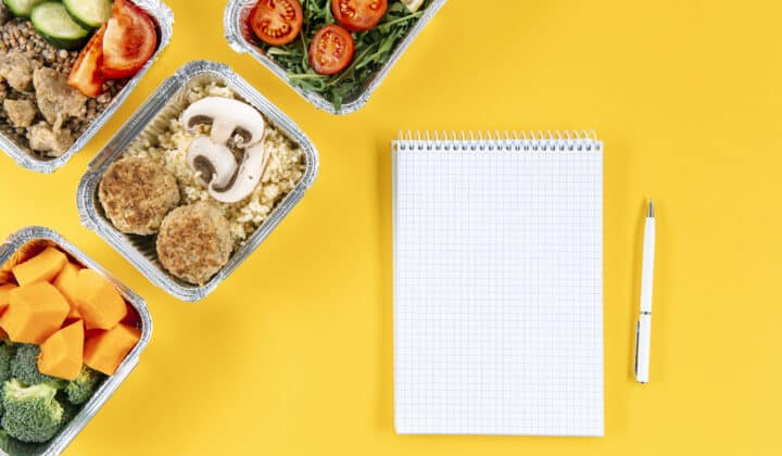 A blank spiral notebook and pen next to foil containers of cooked meal prep food on a yellow background