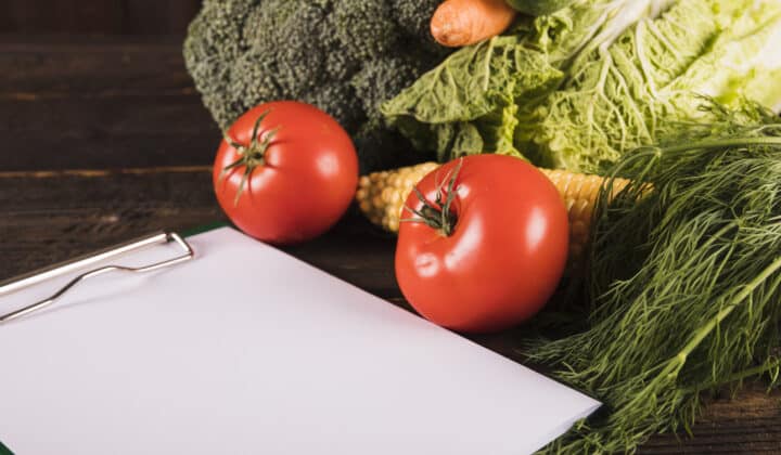 A blank clipboard surrounded by fresh raw vegetables, ready for a budget grocery list