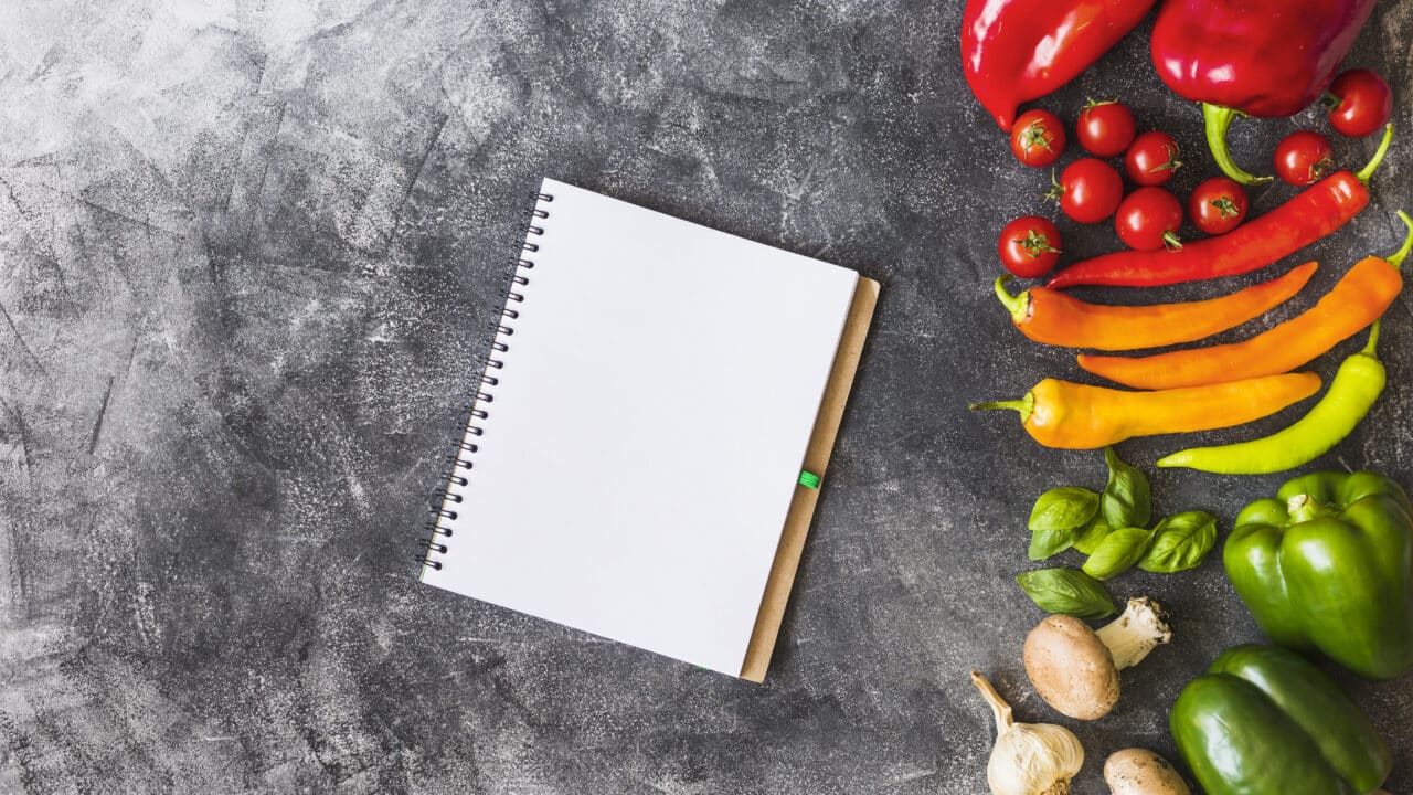 Carnet vierge posé près de poivrons, tomates cerises, piments, champignons et ail frais sur un plan de travail gris.