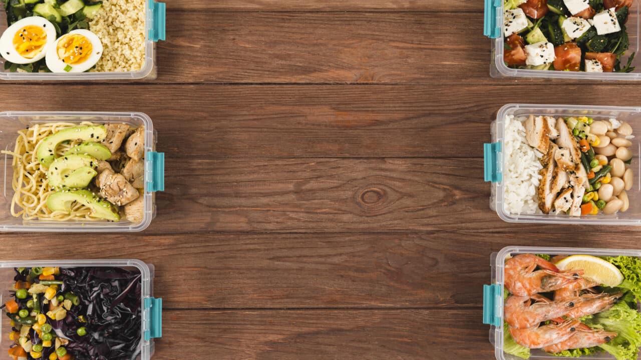 Six plastic meal prep containers with eggs, chicken rice, noodles, shrimp, and vegetable sides on a wood table