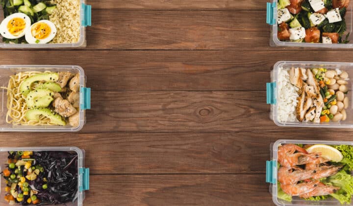 Six plastic meal prep containers with eggs, chicken rice, noodles, shrimp, and vegetable sides on a wood table