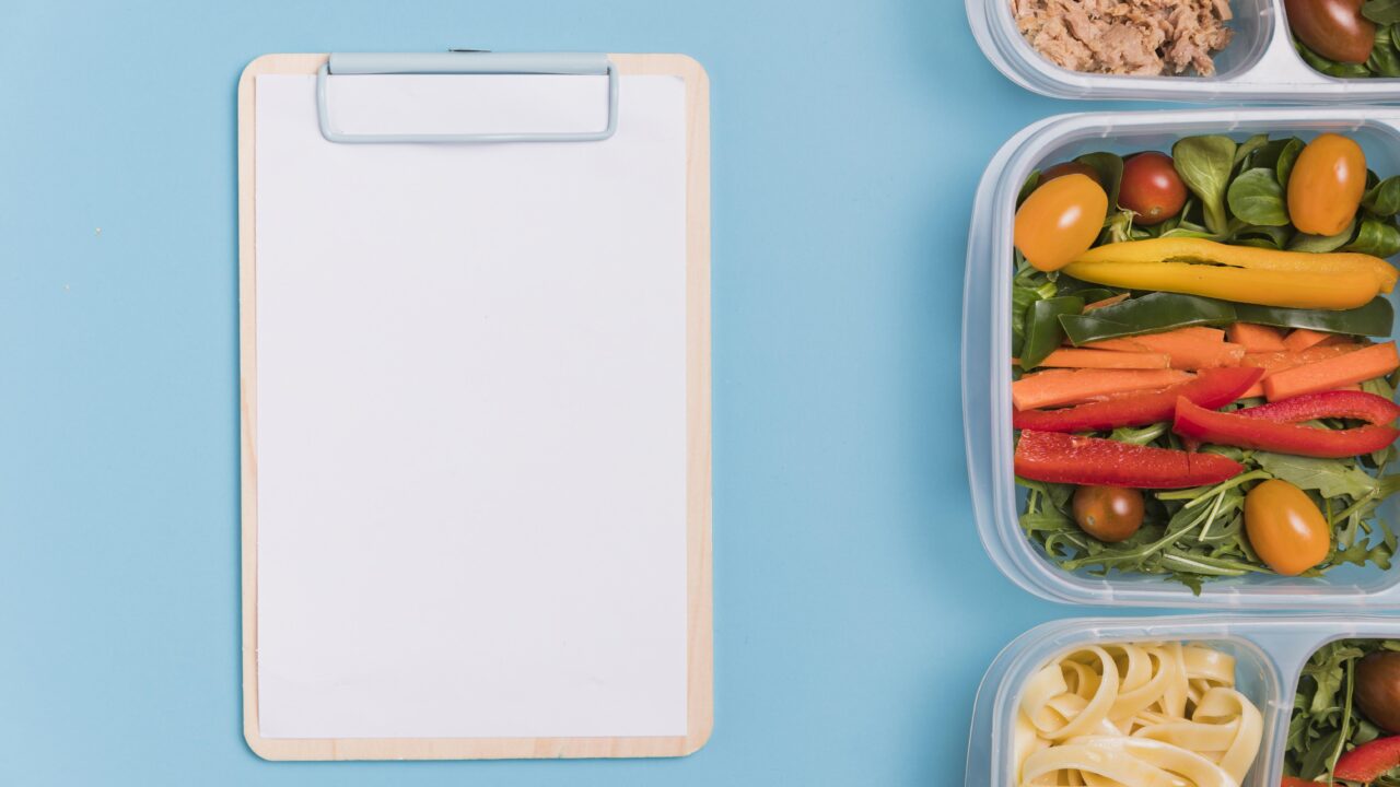 A blank clipboard next to open meal prep containers of tuna, salad vegetables, and pasta on a blue background