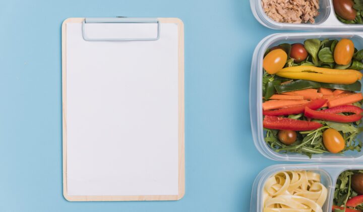 A blank clipboard next to open meal prep containers of tuna, salad vegetables, and pasta on a blue background