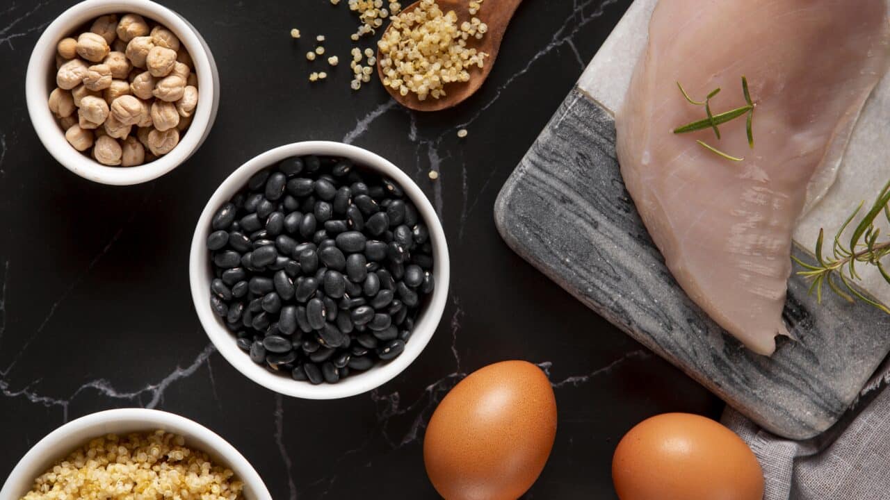 Raw high-protein ingredients for meal prep — black beans, chickpeas, quinoa, a raw chicken breast, and whole eggs arranged on a dark surface