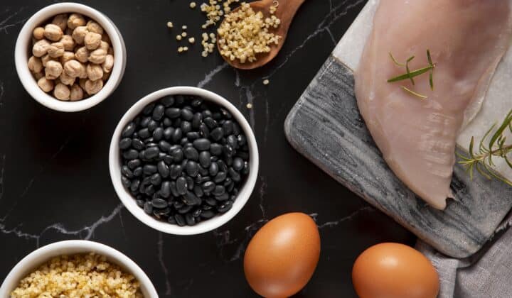 Raw high-protein ingredients for meal prep — black beans, chickpeas, quinoa, a raw chicken breast, and whole eggs arranged on a dark surface