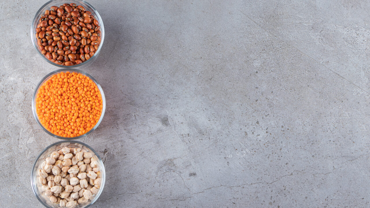 Three glass bowls of dried kidney beans, red lentils, and chickpeas on a gray stone countertop