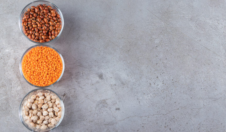 Three glass bowls of dried kidney beans, red lentils, and chickpeas on a gray stone countertop
